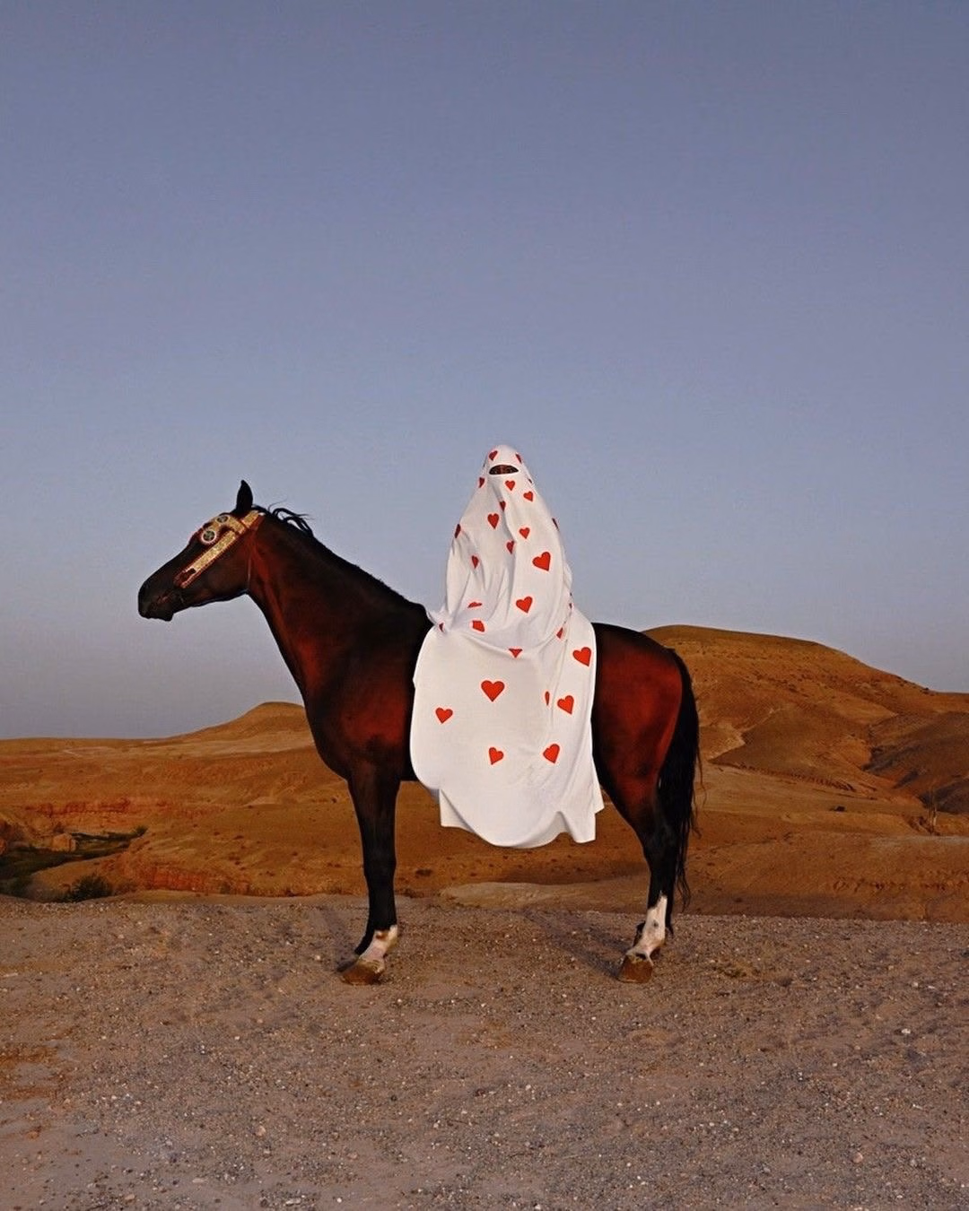A rider in a white cloak covered in red hearts on a dark horse, desert hills at dusk